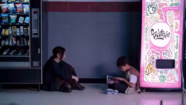 A young woman sits beside a vending machine as a boy reads on the floor