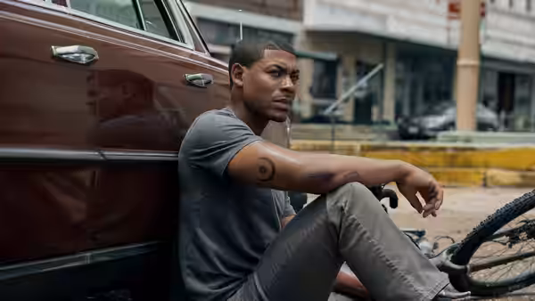 Terry Richmond (Aaron Pierre) sits beside a parked car near a bicycle