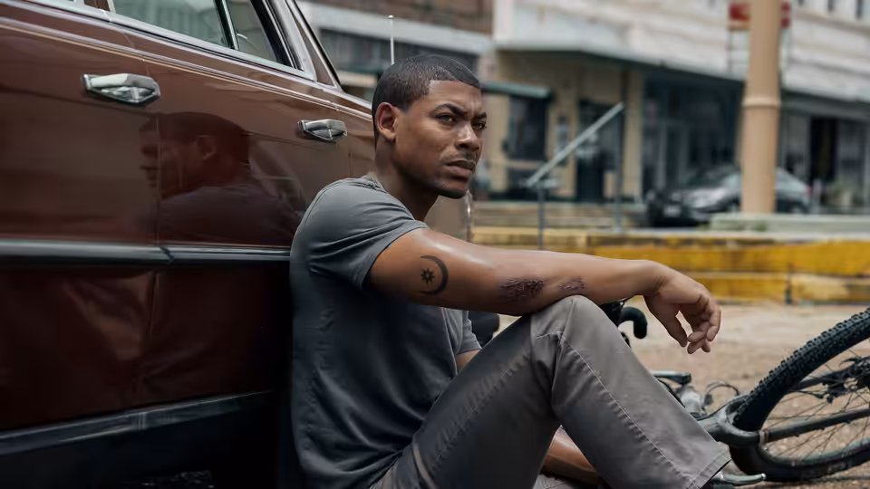 Terry Richmond (Aaron Pierre) sits beside a parked car near a bicycle