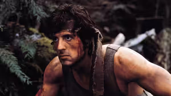 John Rambo (Sylvester Stallone) crouches among rocks and ferns in the woods