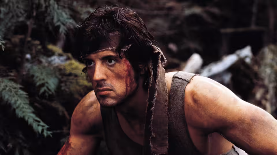 John Rambo (Sylvester Stallone) crouches among rocks and ferns in the woods