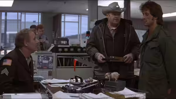 John Rambo (Sylvester Stallone) stands in a police station as officers gather around a desk