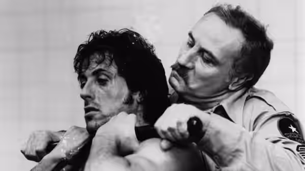 A uniformed officer restrains John Rambo (Sylvester Stallone) from behind during his arrest