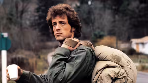 John Rambo (Sylvester Stallone) carries a bedroll and holds out a cup beside the road
