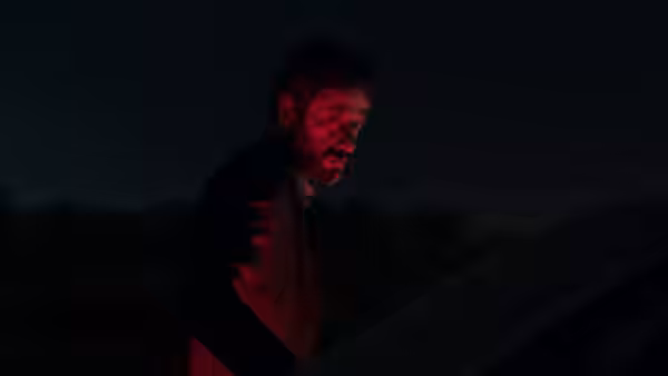 A man leans beside a vehicle, his face lit red in the darkness