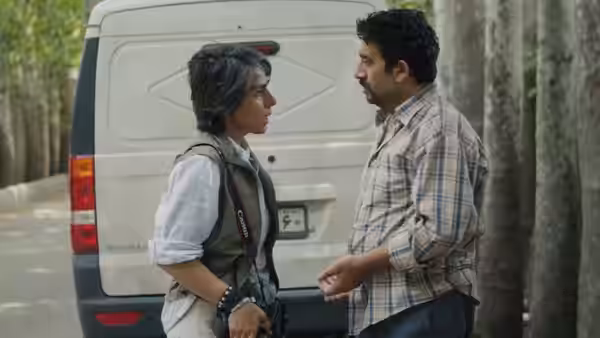 A woman and a curly-haired man talk beside a van among the trees