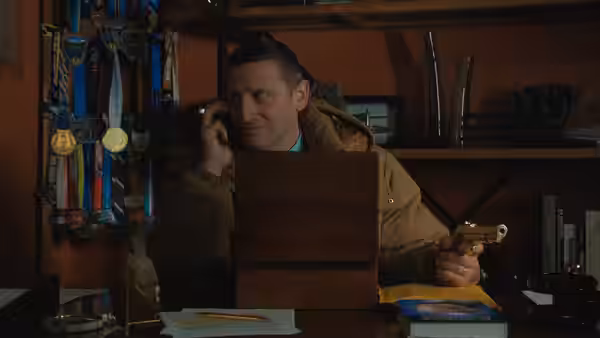 Craig Waterman (Tim Robinson) talks on the phone behind a wooden box, holding a small pistol