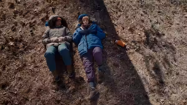 Two women lie side by side on dry grass, one wearing a blue jacket
