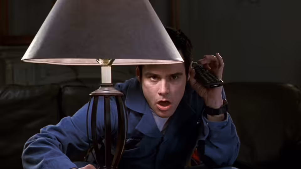 Cable Guy (Jim Carrey) crouches behind a lamp, holding a remote to his ear