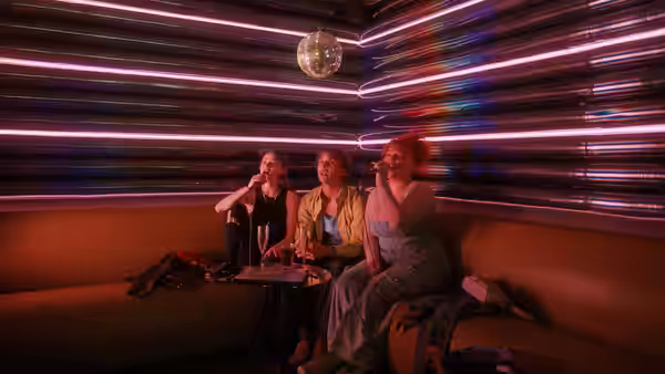 Three women sit together in a neon-lit lounge beneath a disco ball