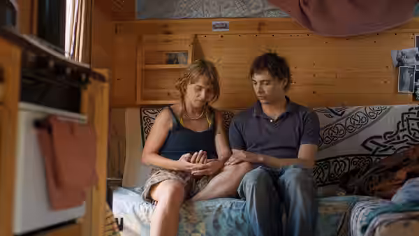 Mike (Frank Dillane) and a woman sit side by side on a sofa, holding hands