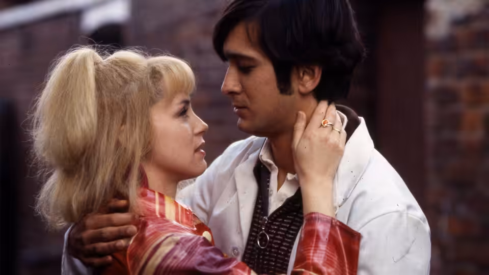 A blonde woman and dark-haired man embrace and gaze at each other