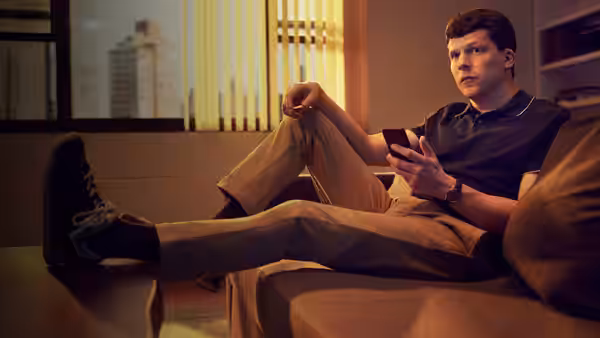 Toby (Jesse Eisenberg) lounges on a sofa, holding his phone