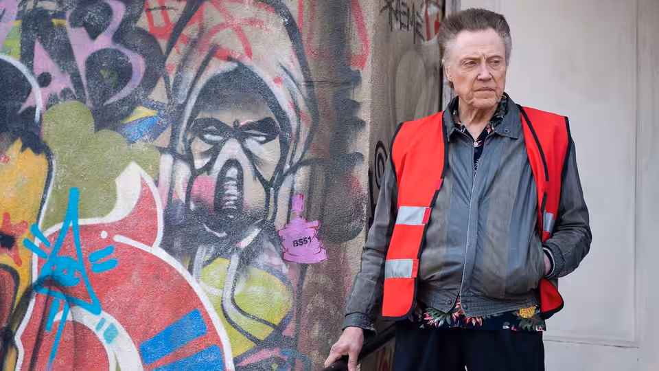 Frank (Christopher Walken) stands beside a graffiti-covered wall in a red reflective vest