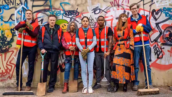 Volunteers stand with brooms and shovels before a graffiti-covered wall