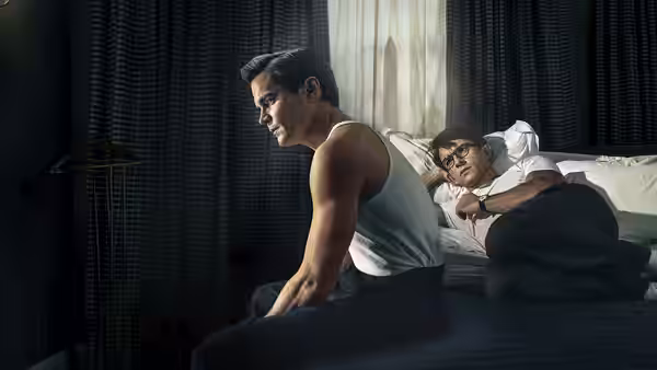 Hawk Fuller (Matt Bomer) sits beside Tim Laughlin (Jonathan Bailey), who lies in bed wearing glasses