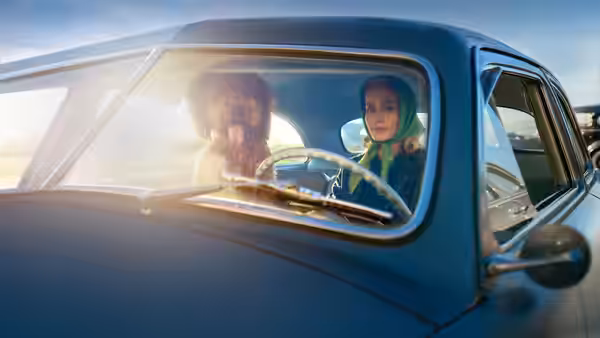 A woman in a green headscarf rides beside a shaggy dog in a blue car