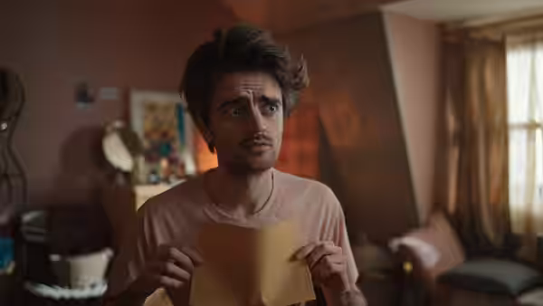 Kash (Bilal Hasna) holds a yellow sheet of paper in a living room
