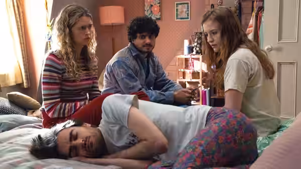 Carrie, Kash, and Jen sit beside Jizzlord, who lies curled on a bedroom bed