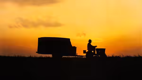 A man on a tractor pulling a trailer silhouetted against an orange sunset