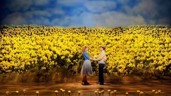 A couple stands holding hands on a wooden stage before a painted flower field