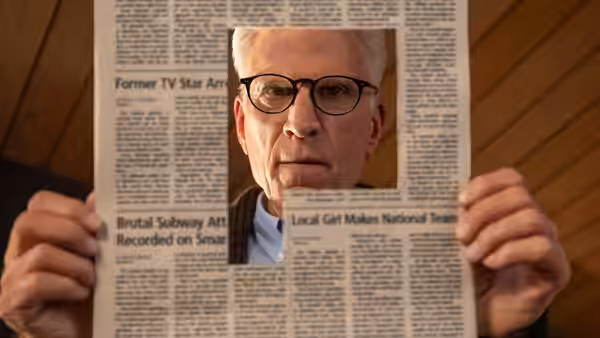 Charles Nieuwendyk (Ted Danson) peers through a hole in a newspaper