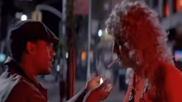 A man in a cap speaks with a curly-haired woman on a neon-lit street