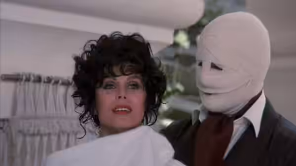 A curly-haired woman stands beside a bandage-wrapped man in a bright room
