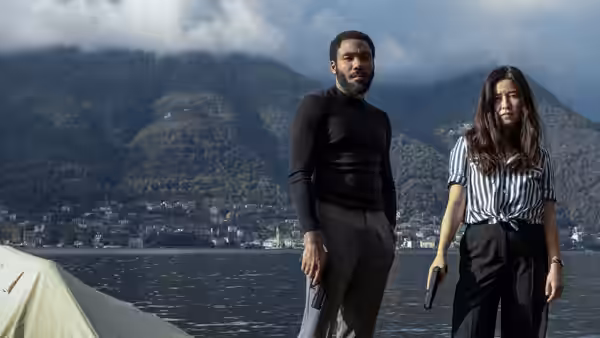 John Smith (Donald Glover) and Jane Smith (Maya Erskine) stand armed beside a lake beneath forested mountains