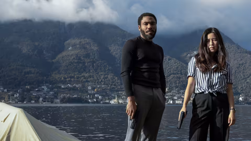 John Smith (Donald Glover) and Jane Smith (Maya Erskine) stand armed beside a lake beneath forested mountains