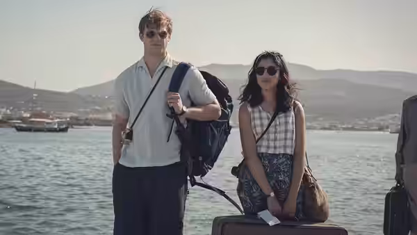 Emma (Ambika Mod) and Dexter (Leo Woodall) wait with luggage beside the water