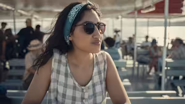 Emma (Ambika Mod) sits at an outdoor waterfront café wearing sunglasses