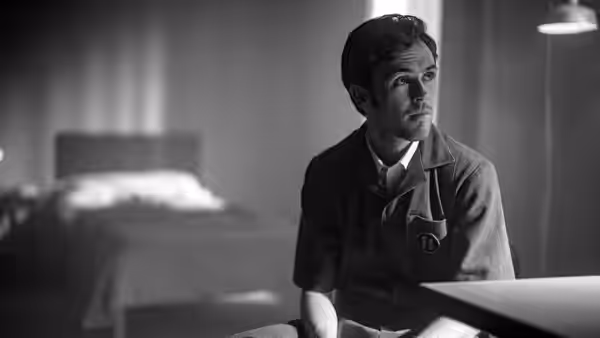 A young man sits alone beside a bed in a stark monochrome room