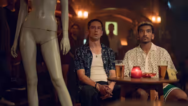 Dionysus (Nabhaan Rizwan) and a companion sit watching a mannequin in a dim bar