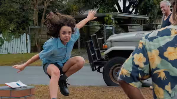 A curly-haired child leaps beside a parked utility vehicle as an older man watches