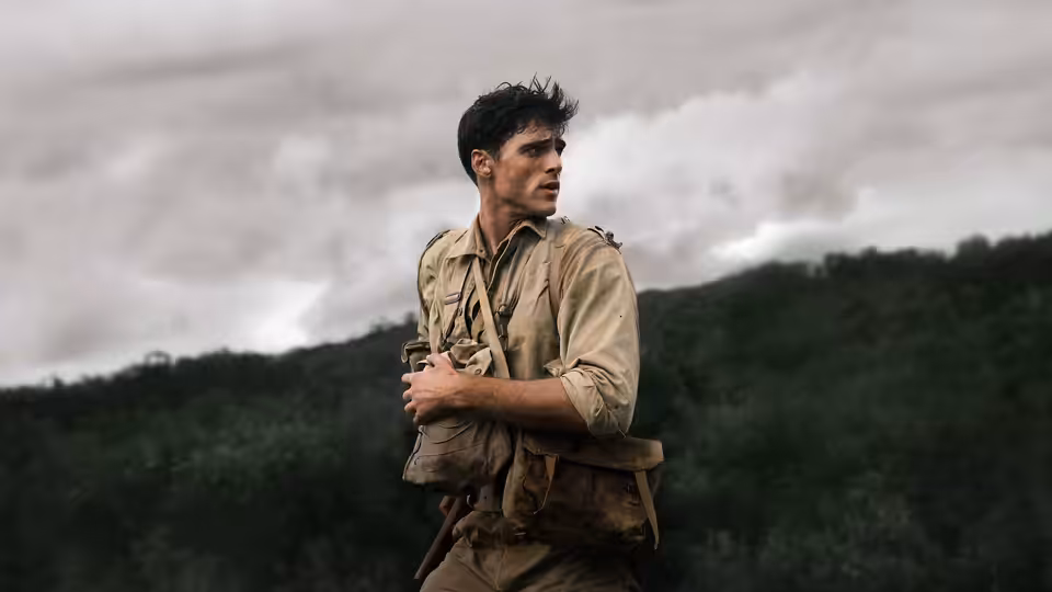 Dorrigo Evans (Jacob Elordi) in khaki uniform carrying a pack across a wooded hillside