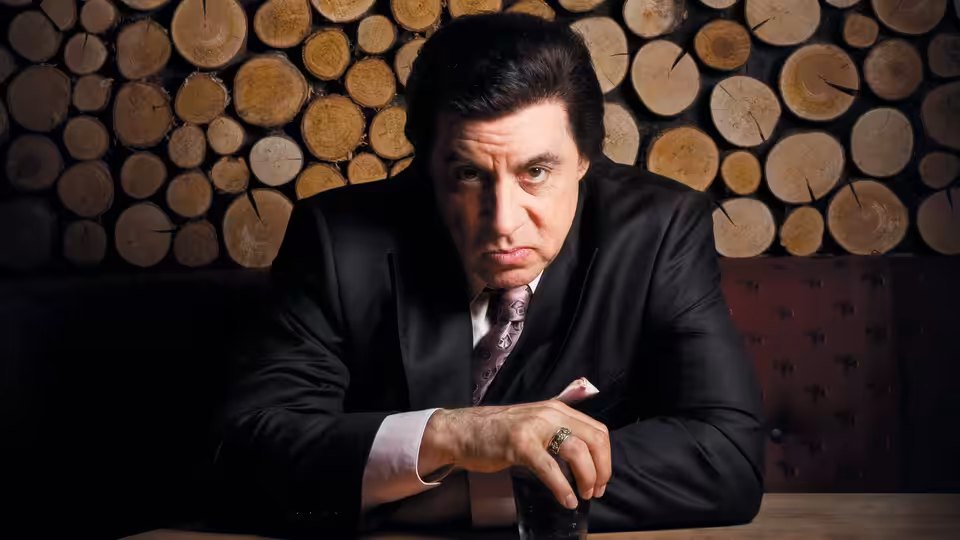 Frank Tagliano (Steven Van Zandt) sits at a table before a wall of stacked logs