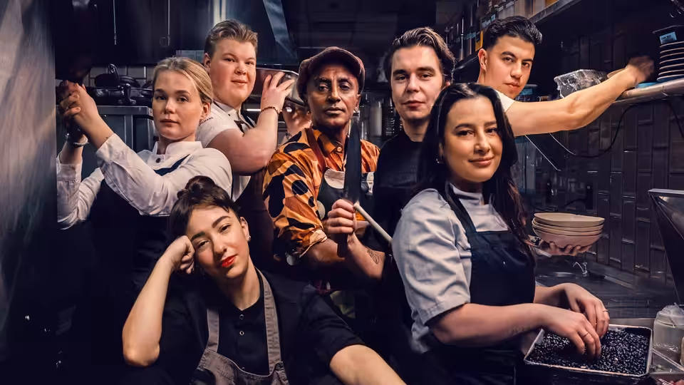 Self (Marcus Samuelsson) and cooks gather around a kitchen counter as one stirs a pan