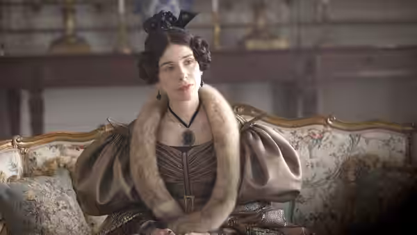 A woman in an elaborate gown sits on an ornate sofa in a drawing room