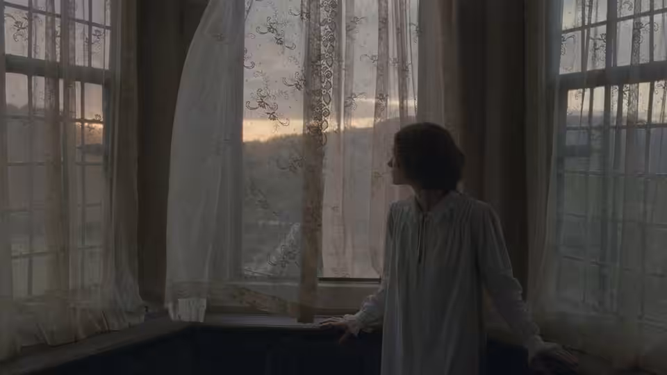 Jane Eyre (Mia Wasikowska) stands by a tall window, looking out over the landscape