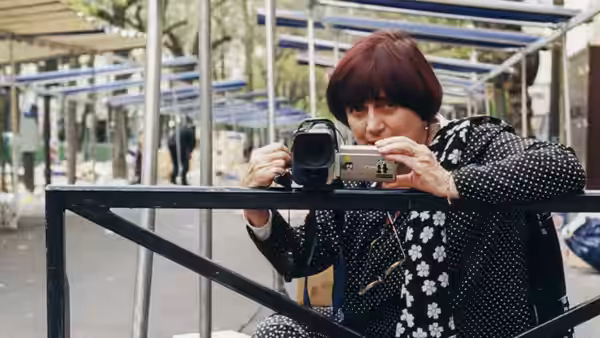 Agnès Varda films through a camcorder behind a metal barrier