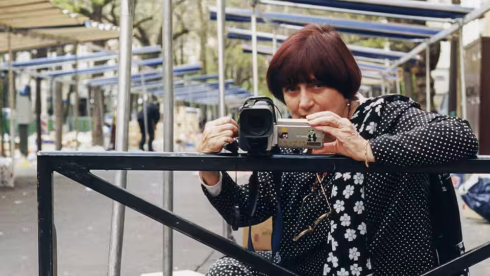 Agnès Varda films through a camcorder behind a metal barrier