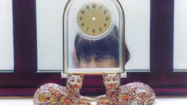 A woman peeks through a clock above two ceramic cat figurines