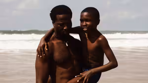 A shirtless man and boy embrace in shallow ocean waves