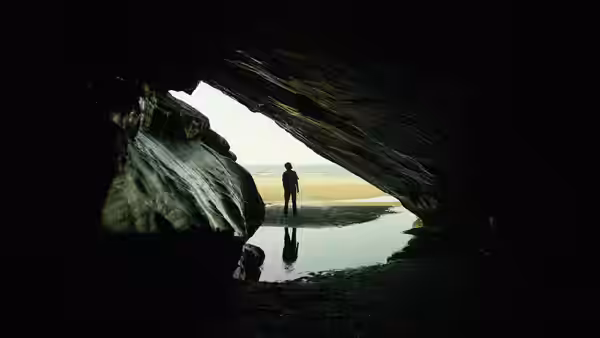 A solitary figure stands at the mouth of a dark cave, facing the shore