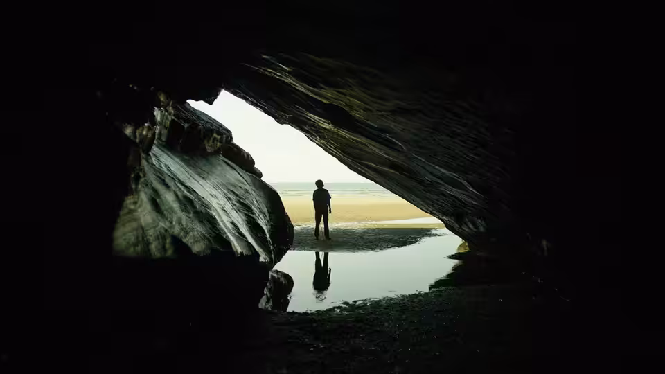 A solitary figure stands at the mouth of a dark cave, facing the shore