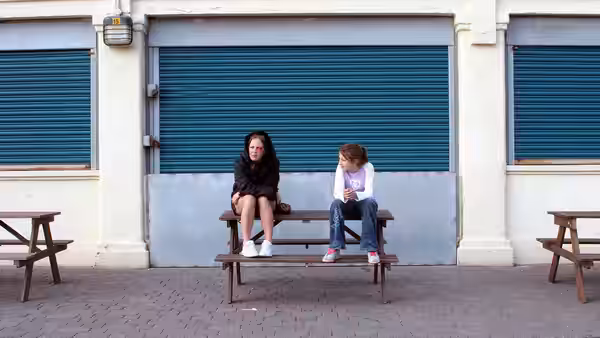 Kelly (Lorraine Stanley) and Joanne (Georgia Groome) sit together on a picnic table outside shuttered shops