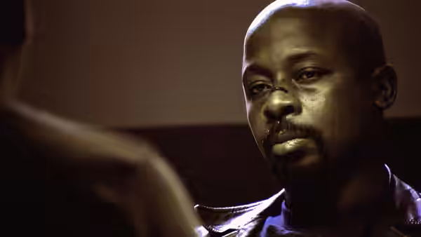 A bald man in a leather jacket faces someone across a dimly lit room