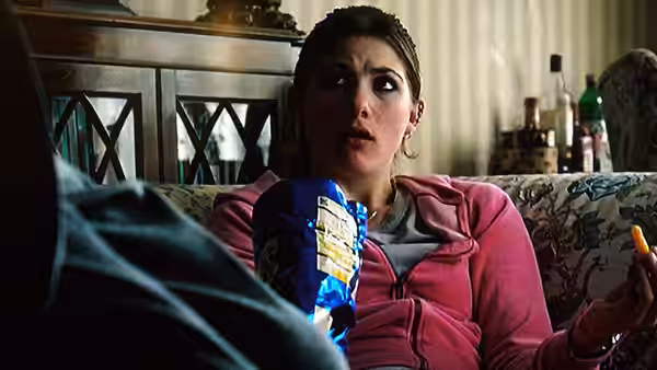 Jessie (Jodie Whittaker) sits on a sofa holding a snack bag and reacting to someone