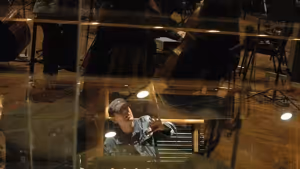 A man works at a recording console behind reflective studio glass